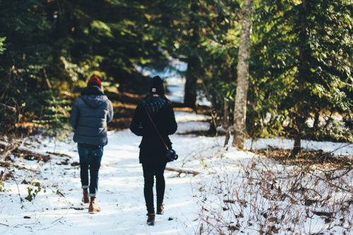 Two people walking through woods with snow on the ground